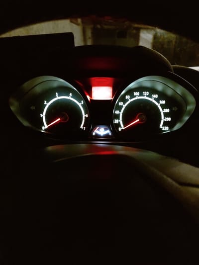 The image shows the illuminated dashboard of a car at night. The dashboard features two main gauges: a tachometer on the left, with a needle pointing to approximately 0.8, and a speedometer on the right, indicating a speed of roughly 20 km/h. There is also a digital fuel gauge with a low fuel warning light illuminated. The background is dark and out of focus, suggesting the car is stationary or moving slowly in a dimly lit area. The setting is identified as Tervel, Bulgaria.