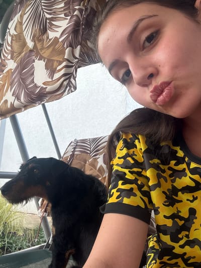 A young woman with dark hair is taking a selfie outdoors, her face close to the camera, puckering her lips. She is wearing a bright yellow shirt with a black camouflage pattern. To her left and slightly behind her, a black dog with brown markings is visible, looking to the left. They are sitting on a swing set with patterned cushions. The background includes foliage and a white wall or sky. The location is Bulgarovo, Bulgaria.