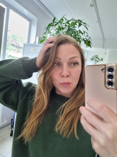 The image is a selfie taken from a low angle, with the camera held by a person's hand. The subject is a woman with fair skin, freckles, and blonde hair. She is wearing a dark green sweatshirt. Her left hand is touching her hair, and her expression is relaxed. The background shows an indoor setting, with a window and some greenery from a houseplant. The time of day appears to be daytime, with natural light coming from the window. The overall mood is casual and personal. There is no visible text in the image.
