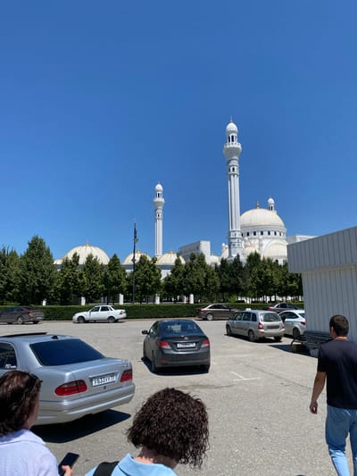 The image shows the "Heart of Chechnya" Mosque in the city of Shali, Russia, on a clear, sunny day. The mosque is a large, white structure with a prominent dome and a tall minaret. Several cars are parked in a lot in front of the mosque, and a few people are visible in the foreground, walking or standing. Trees line the perimeter of the mosque grounds, and a white building is situated to the right of the frame. The sky is a vibrant blue.