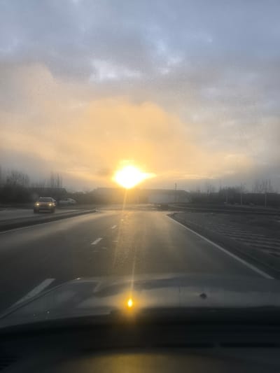 The image displays a forward-facing perspective from inside a vehicle on a roadway in Pakhtakor, Uzbekistan. A low, intense golden sun, partially obscured by clouds, is positioned in the upper center-left, illuminating the sky with warm golden-orange tones that transition to light grey-blue at higher altitudes. The asphalt road surface appears wet and reflective, featuring white solid and broken lane markings. A light-colored SUV, with illuminated headlights, is visible in the distant left lane, moving towards the viewer. Another lighter-colored vehicle is stationary further back on the left side of the road. The distant horizon includes several low-rise buildings and bare trees. The foreground shows the upper portion of a dark vehicle dashboard, reflecting the sun as a bright golden point, with a small dark blemish visible to its right. The overall lighting indicates either sunrise or sunset conditions.