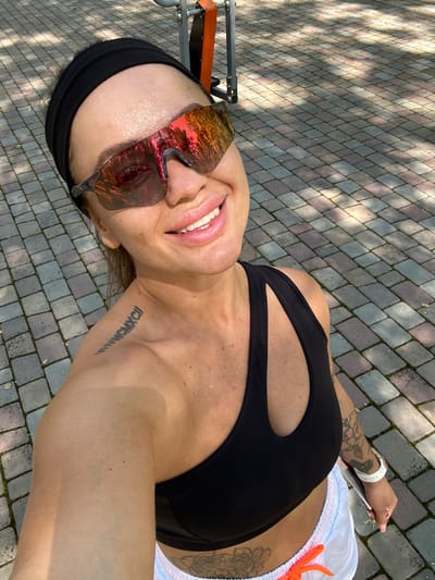 A woman is taking a selfie outdoors in Duvankul', Russia. She is wearing a black headband, mirrored sunglasses, and a black sports bra. White shorts with a pink and blue trim are visible at the bottom of the frame. The woman has a tattoo on her left shoulder and arm, and another tattoo on her upper thigh. The ground is paved with interlocking stones, and shadows from trees are cast across the pavement. A piece of exercise equipment with orange accents is visible in the background. The lighting suggests it is daytime, likely sunny.