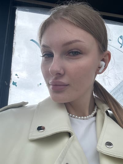 The image is a tilted, close-up portrait of a young woman, taken indoors looking out through a window. The woman has fair skin, light brown hair styled into a ponytail, and light eyes, gazing directly towards the camera with a neutral, soft expression.
She is wearing a white t-shirt, a single strand pearl necklace, and a cream or off-white leather-look jacket with a visible silver snap button on the collar. A white wireless earbud is in her right ear.
The background seen through the window is a bright, uniformly white or light gray sky, indicative of an overcast or cloudy daytime. A dark window frame is visible along the bottom and left edges of the image. Faint, blurry blue shapes are discernible through the window, possibly reflections or small markings on the glass.
The photograph captures a candid moment, with the woman positioned against the bright backdrop of the window. No discernible text is visible in the image. The location context for this photo is Кондратово, Russia.