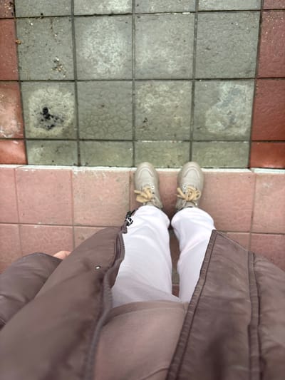 The image, taken from an overhead perspective, shows a person standing on a tiled surface. The tiles are a mix of grey and reddish-pink. The person is wearing white pants and light-colored sneakers with tan laces. A brown puffer jacket or vest is partially visible, obscuring part of the view. The location is identified as Shul'gino, Russia. The image mainly captures the person's footwear and the ground, with minimal surrounding context.
