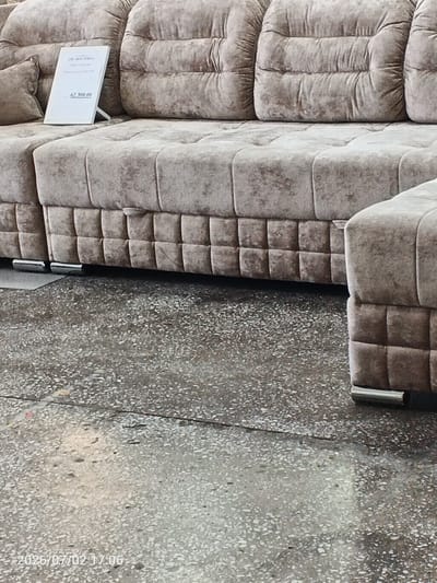 A plush, beige sectional sofa is displayed on a concrete floor in what appears to be a furniture store. The sofa features a tufted design with deep channels on the back cushions and seat. A small white sign is propped up on the sofa's seat, displaying text and a price. The visible text on the sign reads "62 300.00". The floor is a speckled grey concrete, showing some wear and tear, with a faint reflection of light on its surface. The lighting suggests an indoor setting, possibly during daylight hours, but the exact time of day is not clear. The scene is static, with no people or animals present, and no discernible activity is taking place. The overall impression is one of a product display, showcasing a piece of furniture for potential customers. The date stamp on the image indicates July 2, 2026, at 17:06. The location context is Chany, Russia.