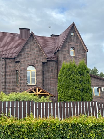 The image shows a brown brick house with a brown tiled roof, a chimney, and several windows. In front of the house, there is a brown metal fence with a green hedge behind it. There are also several evergreen trees and bushes around the property. The sky is overcast. This is a scene in the city of Shchomyslitski Sel'ski Savet, Belarus.