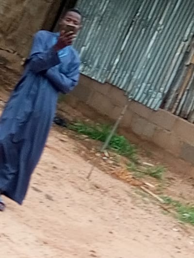 In the city of Wuro Dole, Nigeria, a young Black male, appearing to be in his late teens or early twenties, stands outdoors, holding a smartphone and seemingly capturing a selfie. He is dressed in a traditional blue robe, possibly a kaftan or agbada, which drapes over his body. The background features a textured wall constructed from reddish-brown bricks, with a section of corrugated metal sheeting overhead, suggesting a somewhat rustic or unfinished structure. Sparse patches of green grass are visible near the base of the wall, interspersed with dirt and debris. The ground in the foreground is predominantly sandy or dusty. The overall impression is a casual, candid moment captured in a simple, outdoor setting.