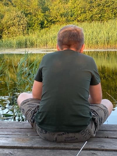 A man is sitting on a wooden dock, facing away from the camera. He is wearing a dark green t-shirt and camouflage shorts. The shorts have text printed on them, including "TOP STAR", "SOUNDS VISION", and "FASHION CHA". He appears to be fishing, with a fishing rod extending into the water. The setting is natural, with tall grasses and trees on the left side of the image, and calm water on the right. The water reflects the surrounding greenery and the sky, suggesting it's daytime with clear weather. The man's posture indicates relaxation and concentration on his activity. The wooden dock is weathered, with visible planks and nails. The overall scene conveys a sense of peaceful outdoor recreation.