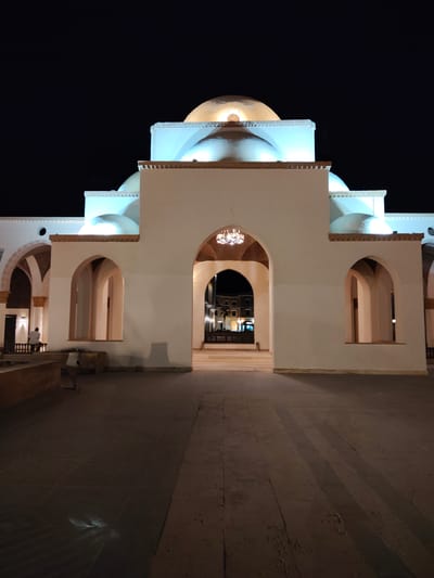 The image shows the exterior of a white building with multiple domes, illuminated at night. The architecture features large arched openings and decorative elements on the upper levels. In the foreground, a paved courtyard with geometric patterns is visible, with some low walls and planters. A single person is seated on a low wall to the left, facing away from the camera. The background shows more of the building's structure, with arches leading into darker interiors where lights are visible. The overall scene suggests a public building or complex, possibly a mosque or a cultural center, at night. The lighting creates a dramatic contrast between the brightly lit white structure and the dark night sky.
