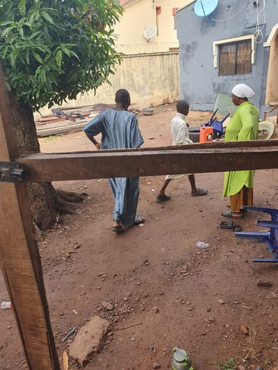 This media file captures an outdoor scene, likely an open yard or pathway between buildings, with a reddish-brown dirt ground. Three individuals are present. One person, on the right, wears a white head covering and a bright green long-sleeved top with a patterned skirt, and black sandals. Another individual, closer to the center, is dressed in a light-colored (possibly white or cream) top and dark trousers, wearing black sandals or slip-on shoes. The third person, on the left and partially obscured by a wooden beam, wears a blue or grey striped tunic and trousers, also with sandals.
The activity appears casual, with individuals standing or moving in the space. The person in green is positioned near blue plastic chairs and an orange jerry can. The person in the light top seems to be facing the person in blue/grey.
Notable details include a light-colored building in the background featuring a dark window and a blue satellite dish with the text "TSTV" visible on it. Another building with a fence topped with barbed wire is also visible. A large, leafy green tree is on the left. The foreground is dominated by prominent wooden beams or posts. Scattered on the dirt ground are various items such as small stones, debris, a plastic bag, a blue plastic spoon, and a plastic bottle with "AQUA" and "WATER" partially visible. The scene is illuminated by natural daylight. The overall setting, clothing, and architectural elements are consistent with Idah, Nigeria. Emotions are not discernible due to the distance and lack of clear facial expressions.