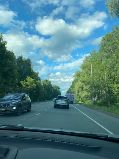 The image is a first-person view from inside a vehicle on a two-lane highway surrounded by dense green trees. The sky is blue with scattered white clouds. In the foreground, the dashboard and windshield wipers of the vehicle are visible. On the highway, there are several cars and a truck with "OZON" branding. A dark-colored SUV is in the left lane, and a silver car is in the right lane ahead of it. The truck is further ahead in the right lane. The license plate of the silver car reads "H775HH 156". The license plate of the dark SUV reads "3EP 716". The scene appears to be a daytime drive through a tree-lined road. Based on the license plate "156", the location is likely within the Tatarstan region of Russia, which includes the city of Zelenodolsk.