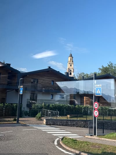 The image shows a street scene in Cavalese, Italy, under a clear blue sky with a few wispy clouds. In the foreground, a paved road curves with a pedestrian crossing marked by white stripes. To the right of the road, a grassy verge is visible. Further back, a modern building with a wooden facade and dark roof dominates the view. A pedestrian crossing sign with a blue square and white silhouette is mounted on a pole, accompanied by two circular traffic signs: one indicating a speed limit of 30 and another with a pedestrian silhouette. The building's glass facade reflects the surrounding mountains and sky. In the distance, a tall, slender bell tower with a dark spire rises above the trees, a characteristic feature of many Italian towns. Lush green trees and hedges surround the buildings and fences, contributing to a well-maintained, residential atmosphere.