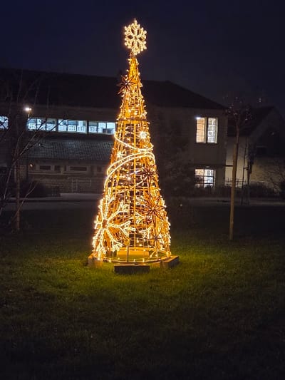 An illuminated Christmas tree sits on a lawn at dusk. The tree is constructed of golden lights arranged in the shape of a traditional evergreen, adorned with snowflake and star motifs. A glowing snowflake tops the tree. Behind the tree is a dark building with lit windows, contrasting the surrounding darkness. The grass is subtly lit by the tree's warm glow, creating a festive ambiance.