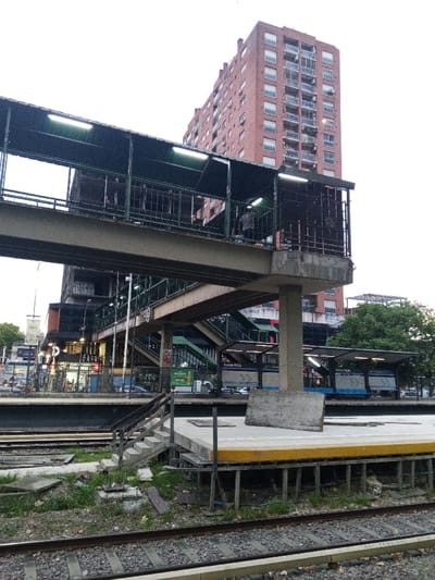 The image depicts an exterior view of the Ramos Mejía train station in Ramos Mejía, Argentina, under an overcast sky. In the foreground, two sets of railway tracks run horizontally across the frame, flanked by a strip of overgrown vegetation and scattered debris. To the right, a concrete train platform features a distinct yellow safety stripe along its edge. A rectangular, flat wooden or concrete board leans diagonally on this platform. Concrete stairs ascend from the track level to the platform on the left.
Dominating the midground is a multi-level pedestrian bridge structure, composed of concrete supports and dark-colored metal framework. The upper walkway is covered by a dark green corrugated roof, beneath which fluorescent lights are illuminated. A single individual, dressed in a dark shirt, is visible on this upper walkway, facing away from the camera. Below this, another bridge section with a similar roof and lighting connects to a series of integrated stairs, leading down towards street level. To the left, beyond the tracks and platforms, ground-level commercial establishments with signs and illuminated storefronts are discernible. On the platform to the right, behind the leaning board, are covered waiting areas equipped with blue benches, and a sign displaying "Ramos Mejía" is visible.
In the background, a tall, multi-story building constructed of reddish-brown bricks with numerous balconies rises against the light grey sky. Green tree foliage is present on the far right. Vehicles are visible on a street beyond the ground-level shops.