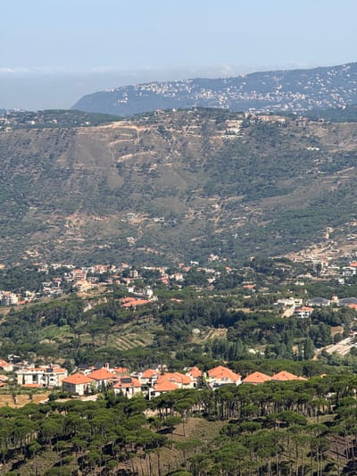 The image is a panoramic view of a mountainous landscape in Mdairej, Lebanon, showcasing a vast expanse of rolling hills covered in dense evergreen trees and dotted with numerous residential buildings. The architecture of the houses is characterized by red-tiled roofs and light-colored walls, interspersed with the vibrant green of the surrounding forest. In the foreground, a dense forest of tall, slender pine trees creates a natural barrier, with their dark green canopy stretching across the lower third of the frame.
As the eye moves upwards, the terrain becomes more rugged, with terraced slopes indicating agricultural activity or cleared land for construction. Patches of scrubby vegetation and exposed earth are visible on the steeper inclines. Further into the distance, the landscape transitions to broader hills and mountains, also sparsely populated with buildings.
The middle ground features a more concentrated cluster of residential buildings, seemingly a small village or a suburban development, nestled among the trees. These houses, with their characteristic red roofs, are built in close proximity, forming a distinct architectural pattern against the natural backdrop.
In the far background, more distant mountains are visible, with a hazy atmosphere obscuring the finer details. A thin layer of clouds hovers near the horizon, suggesting a clear day with potential for atmospheric perspective. The overall impression is one of a serene, semi-rural setting with a significant presence of human habitation integrated into a heavily forested and mountainous environment. No people are discernible in the image. The lighting suggests it is daytime, likely late morning or early afternoon, with the sun casting shadows and highlighting the textures of the landscape.