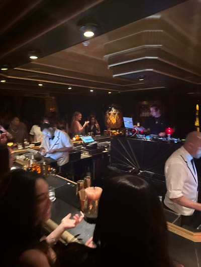 The image depicts a crowded bar scene, likely in the evening, with a focus on a DJ setup and patrons enjoying drinks. The atmosphere appears lively and dimly lit.
People:
Several people are present, including:
- A DJ: positioned behind a mixing console, wearing a white shirt.
- Bar patrons: seated or standing at the bar, some interacting with each other, with drinks in front of them.
Subjects:
- DJ equipment: a mixing console with various controls and screens.
- Drinks: various glasses containing beverages are visible on the bar.
Objects:
- Bar counter: a long, polished bar surface cluttered with drinks and other items.
- Bar stools: chairs for patrons to sit at the bar.
- Lighting: spotlights are embedded in the bar's architecture.
Actions:
- Patrons are ordering and consuming drinks, listening to music.
- The DJ is operating the mixing console, likely playing music.
Interactions:
- Patrons are talking and gesturing to each other.
- There is an interaction between bartenders and patrons.
Environment:
- Interior: The scene appears to be inside a bar or club, with dark, modern décor.
- Lighting: The lighting is dim and atmospheric.
- Location: Based on the style and atmosphere of the image, it is possibly located in Miami Beach, United States.