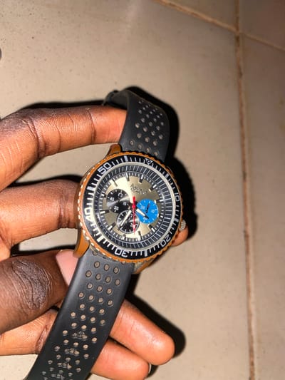 An eye-level close-up shot features a person's dark-skinned hand holding a watch against a light-colored, tiled surface. The setting appears to be indoors, likely in Hayin Banki, Nigeria, with artificial lighting casting a shadow behind the watch.
The main subject is a men's chronograph wristwatch with a distinct design. It has a round, two-tone dial; the center is a metallic gold or bronze, transitioning to a black outer ring with white minute markers. Three chronograph subdials are visible: one blue subdial at the top displaying "10" and "30", and two black subdials below, marked "15, 30, 45" and "20, 40, 60" respectively. A prominent red second hand points towards the "20" marker on the main dial, while the hour and minute hands are white and black. The words "AMERICAN" and "CHRONOGRAPH" are partially visible on the main dial.
The watch features a black rotating bezel with white numbers and markers (10, 20, 30, 40, 50, 60), indicating it might be a dive watch. The watch case and bezel edge show a worn, bronze or copper-like coloration. A silver crown and pushers are seen on the right side of the watch. The watch is attached to a black rubber strap, characterized by a pattern of small circular perforations, with some visible debris or dirt on its surface. The person's fingers are visible, particularly the thumb and index finger, holding the watch securely. The overall impression is one of showcasing or examining the timepiece.