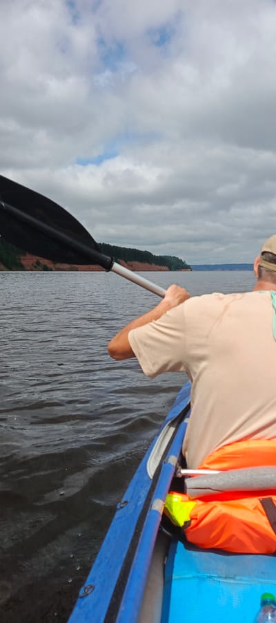 The image shows a person kayaking on a body of water. The person is wearing a tan t-shirt and a baseball cap. They are holding a black and silver paddle, which is partially submerged in the water. In the background, there is a tree-lined shore with a red-brown cliff face. The sky is cloudy. The scene is peaceful and serene. The location is not identifiable as Kostovaty, Russia.