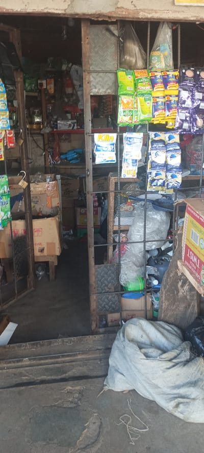 The image depicts a cluttered storefront, likely in a market or commercial area in Kerang, Nigeria. The entrance is partially blocked by a metal gate or bars. Goods are displayed both inside the shop and hanging from the gate, including packets of sweets, coffee, and detergents such as "Klin." Boxes and bags, some appearing to contain merchandise or refuse, are piled near the entrance. The foreground features a large sack and other debris on a concrete surface, suggesting the storefront is located on a street or public walkway. The overall scene conveys a sense of informality and limited space.