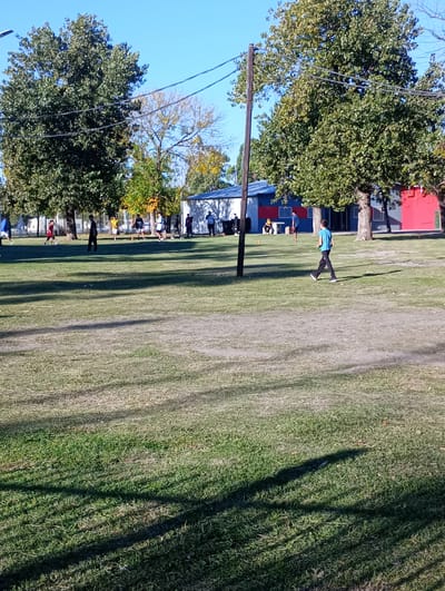 This image depicts an outdoor recreational area, likely a park or sports field, in Villa Domínico, Argentina, bathed in bright sunlight. The scene is dominated by a large expanse of green grass, with several mature trees featuring dense green foliage. Some trees exhibit yellowish leaves, suggesting an autumn season. In the background, simple buildings with blue, red, and white painted exteriors are visible, possibly indicating community facilities or sports club structures. A wooden utility pole carrying power lines stands prominently in the middle-left of the frame.
Numerous individuals, appearing to be children and young adults, are scattered across the grassy area, engaged in various casual outdoor activities. Towards the left, a person in a red shirt is seen near a soccer ball, hinting at a game or practice session. Other people are visible further back near the buildings, some standing and observing. In the mid-ground on the right, a young boy in a light blue t-shirt and dark pants walks leisurely across the grass, carrying a small green object in his hand.
Long, distinct shadows are cast across the grass from the trees and people, indicating that the sun is at an angle, suggesting either late morning or late afternoon. The sky is clear and blue, underscoring the sunny weather. The overall atmosphere appears relaxed and active, focused on outdoor enjoyment and possibly amateur sports. No discernible text is visible in the image.