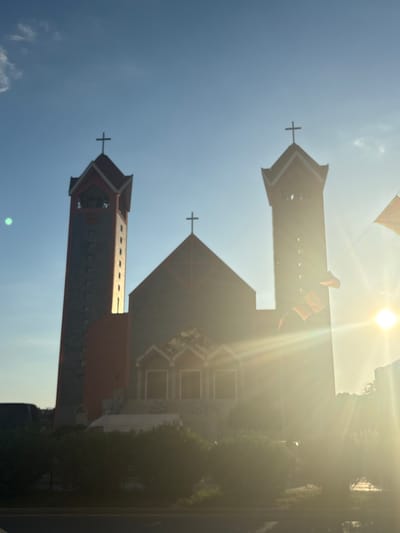 The photograph captures a sun-drenched view of the Church of St. Nicholas in Bjeliši, Montenegro. The church, an Orthodox Christian place of worship, is characterized by its distinct architectural features, including two prominent bell towers flanking a central nave. The taller, left tower is clad in grey stone and features square windows and a clock face. The right tower appears similar in design. Both towers are topped with dark, pointed roofs adorned with crosses. The central part of the church has a distinctive red roof and arched windows. Sunlight streams intensely from the right side of the frame, creating lens flare and obscuring details of the church and its surroundings. Several flags, appearing red and yellow, are visible on poles near the right tower, fluttering in the breeze. In the foreground, lush green bushes obscure the lower portions of the church, and a road is visible at the very bottom of the image. A white van is partially visible behind the bushes. The sky above is a clear, pale blue, with a few wispy clouds on the left side.
