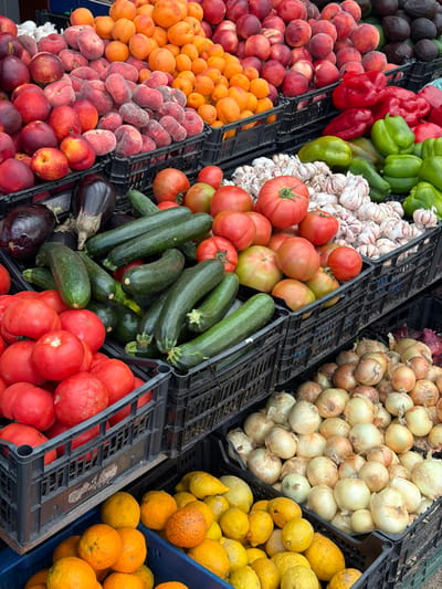 This image depicts a vibrant outdoor market stall laden with a diverse assortment of fresh produce, likely situated in a city like Santiago de Compostela, Spain, known for its bustling markets.
The foreground is dominated by crates overflowing with fruits and vegetables. In the bottom left, a blue crate holds a generous pile of bright orange citrus fruits, possibly oranges or tangerines, some with textured skins. Adjacent to it, to the right, is a crate filled with a mixture of yellow lemons and some larger, similarly colored citrus fruits.
Moving up, a black plastic crate contains a substantial quantity of red tomatoes, their skins smooth and varied in ripeness. Above the tomatoes, another black crate is filled with long, dark green zucchini, some showing slight ridges and visible stems. Interspersed with the zucchini are a few plump, dark purple eggplants.
Further up and to the right, a collection of garlic bulbs with papery white skins are neatly piled. Beside the garlic, large red and green bell peppers add a splash of color. Several large, red tomatoes are also visible in this section.
The upper portion of the image reveals an abundance of stone fruits. Piles of peaches and nectarines in shades of pink, red, and orange are stacked high in black plastic crates. To their right, a large quantity of smaller, bright orange fruits, likely apricots or similar, are visible. In the far upper right corner, dark green avocados can be seen.
The lighting suggests it is daytime, with soft, natural light illuminating the produce. The background is slightly blurred, but suggests further stalls or structures typical of an outdoor market. There are no people visible in the frame, focusing the viewer's attention entirely on the colorful bounty of fresh food. The overall impression is one of abundance, freshness, and the lively atmosphere of a European market.