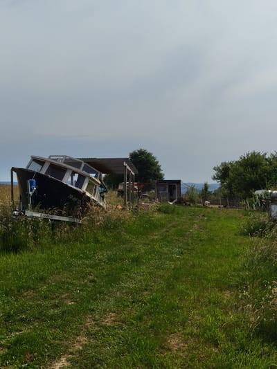 The image shows a dilapidated boat resting on a trailer in a grassy field. The boat is dark in color with white trim around its windows. The text "TR-WF 12" is visible on the side of the boat. Behind the boat, there is a simple wooden structure. Further in the background, trees and a small building can be seen, with a hazy sky above. The location is likely a rural area in or near Mettlach, Germany, given the vegetation and style of the structures, though no specific landmarks are discernible to confirm the exact location within the city.