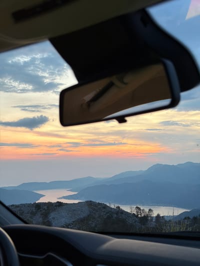 The image captures a view from inside a car looking out through the windshield and rear-view mirror. The scene is a landscape dominated by mountains, a body of water, and a sky filled with clouds. The time of day appears to be either sunrise or sunset, as the sky exhibits hues of orange, pink, and yellow. The mountains in the distance are layered and fade into a hazy blue. The water appears calm, reflecting the sky's colors. The foreground shows the dashboard and steering wheel of the car, indicating the perspective of a driver or passenger. The image is taken in Kuk, Montenegro.
