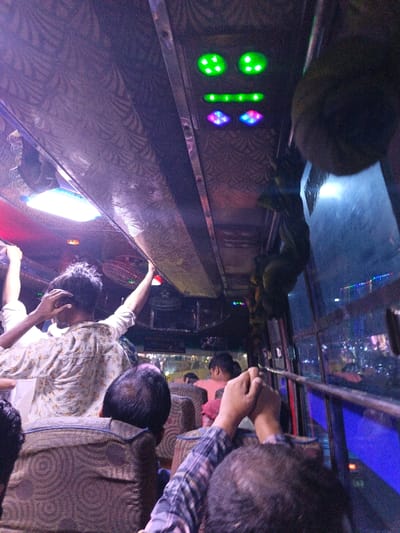 The image depicts the interior of a public transport vehicle, likely a bus, at night in Savar, Bangladesh. The perspective is from a passenger's viewpoint, looking forward and to the right. The ceiling features patterned dark upholstery with an oval fluorescent light fixture, two pairs of circular green LED lights, a horizontal green LED strip, and two diamond-shaped purple/pink LED lights. Two small red-bladed electric fans are mounted overhead.
In the foreground, the top of a balding head is visible, alongside a pair of hands with interlocked fingers belonging to an individual wearing a plaid shirt. Immediately ahead, a person with dark hair, wearing a light-colored patterned shirt, is standing or bracing themselves. This individual has one arm raised and bent near their head, while the other arm extends upward, possibly grasping an overhead bar. Further into the vehicle, additional passengers are visible, some seated in patterned upholstered seats.
Through the window on the right, a dark exterior is seen with blurred city lights, including a prominent blue light reflection. Distant, colorful lights suggest an active urban environment at night. Dark, possibly decorative, hanging elements are visible near the window frame.