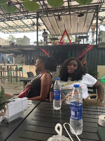Two women sit at a dark wooden table, each with a bottle of Aquafina water. One woman wears a white t-shirt with cartoon characters and has curly hair. The other wears a black tank top with hair styled up. The setting appears to be an outdoor restaurant or bar with a partial roof structure overhead. There are tables and chairs in the background, and fans hang from the ceiling.