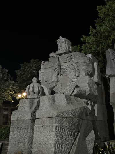 The image displays a large, multi-figure monument constructed from grey, rough-hewn stone or granite, set outdoors at night in Veliki Preslav, Bulgaria. The monument is rendered in an abstract, cubist style. A prominent figure at the top features a blocky, bearded face and a crown, gazing slightly upwards and to the right. To the left of this central figure, a smaller, stylized crown is carved into a separate block. Lower on the monument, another abstract face with a distinctive eye and mouth is discernible, integrated into the textured stone. Artificial lighting illuminates specific sections of the monument, creating strong highlights and deep shadows. In the background, dark green trees are partially visible, with some foliage faintly lit. A street lamp on the left provides additional illumination, and a portion of a dark building with a visible doorway can be seen in the far left background.