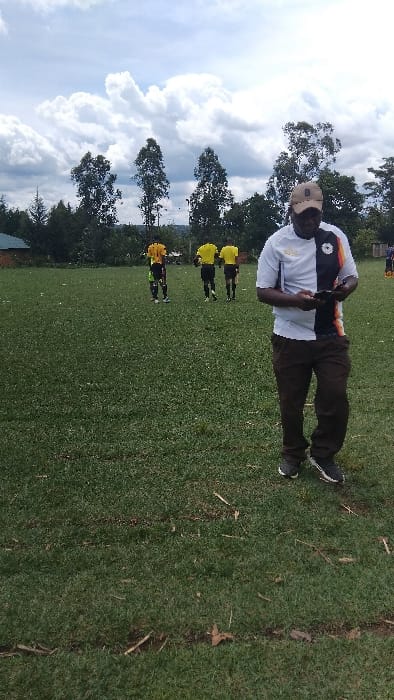 The image shows a man standing on a grassy field in Bungoma, Kenya. The field is bordered by trees and a building in the distance. The man is wearing a white and black shirt with a gold trim, black pants, and a brown baseball cap. He is holding a black cell phone in his hand. There are four men in yellow shirts and black shorts walking in the distance on the field. The sky is cloudy.