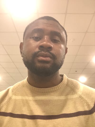 The image displays a head-and-upper-torso shot of an adult male. He has dark skin, short dark hair, and a full dark beard and mustache. His gaze is directed forward towards the camera, and his expression is neutral. He is wearing a long-sleeved, crew-neck knit sweater featuring horizontal stripes; the primary color is beige or light brown, accented with thinner light cream stripes and wider dark brown stripes. The background consists of a light-colored acoustic tile ceiling with a grid pattern and several circular recessed light fixtures, indicating an indoor setting. The specific location in the city of النعيلة, Saudi Arabia, cannot be determined from the visual evidence.