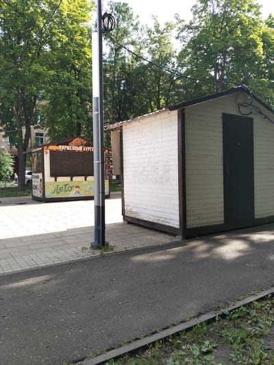 This media file captures an outdoor scene in Korolyov, Russia, on a bright, sunny day. The focus is on two small kiosks situated on a paved walkway, with a street or road in the foreground. Lush green trees form a canopy in the background, suggesting a park or a tree-lined street.

The kiosk on the left is brightly decorated with graphics, advertising food items. Text visible on this kiosk includes "ФИРМЕННЫЙ БУРГЕР" (signature burger), "ПОНЧИКИ" (donuts), and "КОФЕ" (coffee). A large graphic with the word "ЛЕТО" (summer) and images of children and a kite is prominent on its side. The kiosk appears to be open for business, though no people are actively engaged with it at this moment.

The kiosk to the right is simpler, with white wooden walls and a dark door. It seems less decorated and possibly closed. A lamppost stands between the two kiosks, with visible wires and a light fixture.

In the background, behind the kiosks and trees, glimpses of buildings can be seen. The lighting suggests it is daytime, likely late morning or early afternoon, given the strong sunlight casting shadows. The overall mood is relaxed and casual, typical of a pleasant summer day in an urban or suburban setting. The paved walkway is laid with rectangular tiles, and the road in the foreground is asphalt, bordered by grass and vegetation.