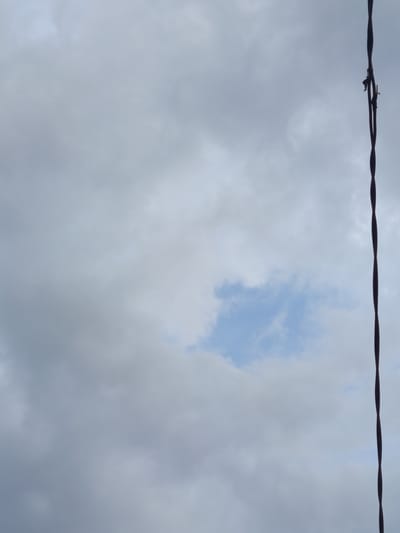The image shows a cloudy sky with a thin, dark wire running vertically along the right side. The wire appears to be barbed, with a small projection visible near the top. A patch of lighter blue sky is visible in the center of the frame, contrasting with the predominantly gray clouds. The image was taken in the city of Жаны-Кыштак, Kyrgyzstan.
