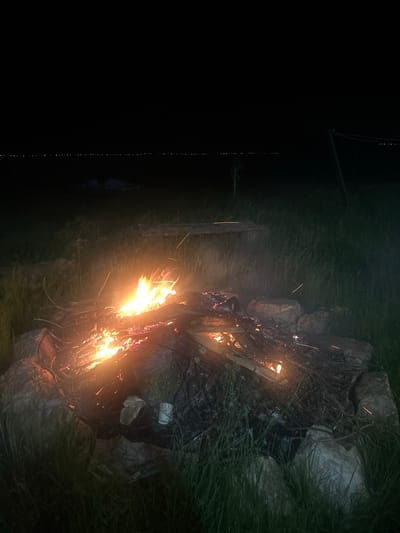 This image depicts a nighttime outdoor scene in Цигов чарк, Bulgaria. The central subject is a campfire contained within a ring of large, irregular rocks, occupying the lower-center of the frame. The fire actively burns with bright orange and yellow flames, consuming wood logs and branches, and displaying numerous glowing red embers. Multiple small, linear sparks are visible, arcing upwards and outwards from the fire. Tall, dark green grass surrounds the campfire and extends into the foreground and midground. To the immediate right of the fire, near the rock ring, a white cylindrical object (possibly a cup) and some lighter-colored debris are visible on the ground. In the midground, behind the fire, indistinct dark shapes suggest additional tall grass and rudimentary wooden structures like planks or benches. Towards the far right, a dark vertical pole with horizontal lines, likely a utility pole or support structure, is visible. The upper portion of the image reveals a completely dark night sky. Along the distant horizon, a distinct, linear series of small, bright white lights is present, indicating a remote settlement or feature. No individuals are depicted.