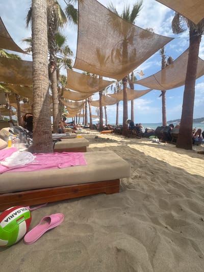 A wide shot captures a sandy beach scene at a resort in Keramoti, Greece. The foreground is dominated by a sun lounger with a pink towel draped over its edge, and a pink flip-flop and a volleyball are on the sand next to it. Beyond this, rows of sun loungers and people are visible under canopies supported by palm trees. The background features the calm sea and distant hills, under a partially cloudy sky. The overall impression is one of a relaxed beach vacation setting.