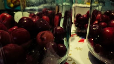 The image shows two glass jars filled with dark red cherries. The jars are positioned on a table with a white tablecloth that has a snowflake pattern. The lighting is dim and the focus is on the cherries within the jars, with some of the cherries appearing to have water droplets on them. The background is out of focus, but hints at a kitchen or dining area. There are no people visible in the image. The image is of a still life, focusing on the preserved fruit. There is no information provided to indicate the location in Troițcoe, Moldova.