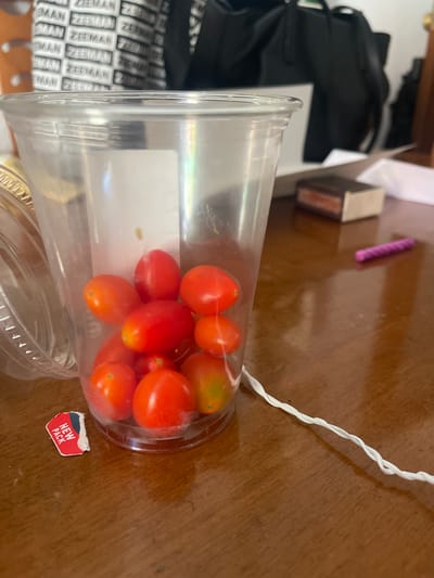 A close-up, eye-level shot shows a clear plastic cup filled with cherry tomatoes sitting on a wooden table. A small, red and white sticker that says "NEW PACK" is adhered to the outside of the cup near the bottom. In the background, out of focus, are a black bag and a stack of items with the word "ZEEMAN" printed repeatedly. A thin, white, twisted cord stretches from the cup across the table. A small, rectangular object, possibly a book or a box, and a thin, purple, cylindrical object are also visible on the table in the background. There are no people present in the image. The scene appears to be indoors, with the lighting suggesting a daytime setting. There is no information in the image that would allow for a determination of its location within Santa Coloma de Gramenet, Spain.
