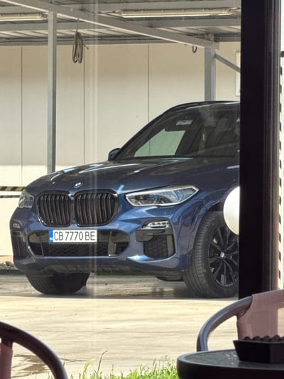 A dark blue BMW car is captured in an outdoor setting, viewed through a reflective window or glass door. The car is positioned with its front facing the left side of the frame, showcasing its prominent black kidney grilles, modern headlights, and a visible BMW emblem. One of its black alloy wheels is partially visible on the right side of the car. The vehicle is parked on a light-colored paved surface, with some green grass or foliage faintly visible on the far right.
The background consists of a light-colored building wall and metal structural elements, suggesting a parking area, possibly a garage or car wash facility. The presence of a dark frame at the top and bottom edges (when the image is viewed in its correct orientation, not rotated) and subtle reflections indicate the photo was taken from inside a building. A brown chair armrest is visible in the foreground on the far right.
The time of day appears to be sunny daytime, with bright light casting some reflections and illuminating the scene.
Visible text includes the Bulgarian license plate "CB 7770 BE" with a small "BG" country code sticker on its left side, indicating the car's origin. Given the context, the location is Elhovo, Bulgaria.