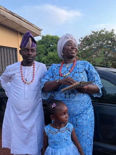 Three people pose together in an outdoor setting in Rafin Bauna, Nigeria. An adult woman wearing a light blue lace dress, a light-colored head wrap, and a red beaded necklace smiles broadly. She holds a brown wallet or clutch in her hands. A young girl wearing a matching light blue lace dress with a pink hair clip stands beside her. An adult man, wearing a white traditional shirt and a purple-striped cap, stands near them, also wearing a red beaded necklace. They stand in front of a dark-colored car, with trees visible in the background. A building is partially seen in the lower left corner. The sky above is light blue with some clouds.