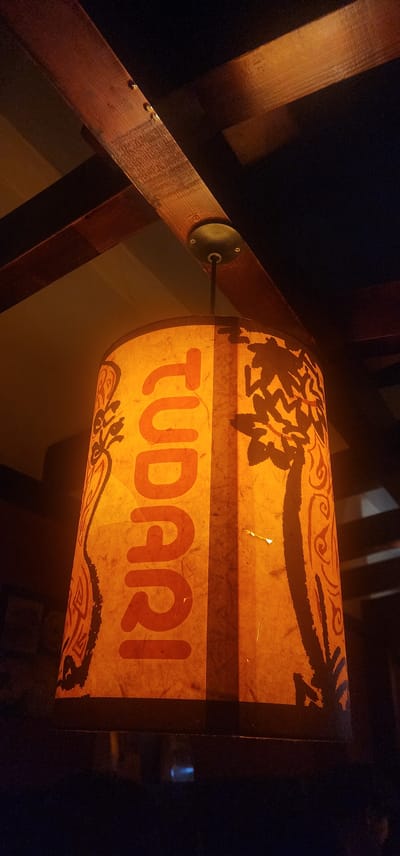 A cylindrical lampshade with a warm orange hue hangs from a ceiling featuring wooden beams. The lampshade is adorned with a graphic design that includes the word "JUDA" in large, bold red letters on one section and a silhouette of a tree with swirling patterns on another. The background is dark, suggesting an indoor setting, possibly a restaurant or cafe, given the ambient lighting and the style of the lampshade and ceiling. The overall impression is one of cozy and artistic lighting.