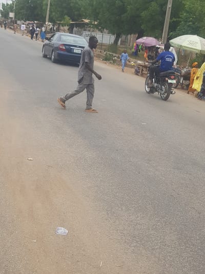 The image is of a street in Dabawa, Nigeria. A man is walking in the street, and a blue car is parked on the side of the road. A motorcycle is parked near a vendor with a blue shirt that has Chinese words on it. Trees and foliage line the left side of the frame, and a group of people can be seen gathered on the side of the road behind the car. The road itself is made of asphalt and has some trash and debris scattered on it.