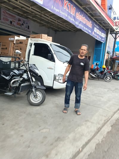 A medium shot shows a man standing in front of a parked white truck loaded with boxes labeled "Panasonic." To the left of the truck is a black three-wheeled motorcycle.  The background includes a storefront with a sign reading "K1M elektronik" and a banner advertising "DEALSEMBER." Two motorcycles are parked behind the truck. The man wears a black t-shirt, rolled-up denim jeans, and sandals.
