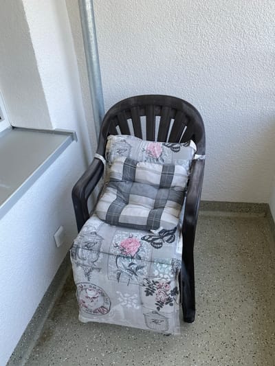 A dark brown plastic chair is positioned on a terrazzo floor, likely on a balcony or patio. The chair is adorned with two cushions. The bottom cushion is a larger seat cushion with a patterned fabric featuring floral designs, birds in cages, and butterflies. The upper cushion is a smaller back cushion with a plaid pattern in shades of gray and white. To the left of the chair, a white textured wall is visible, with a narrow grey shelf extending horizontally. A silver metal pipe runs vertically along the wall behind the chair. The scene is lit by natural light.