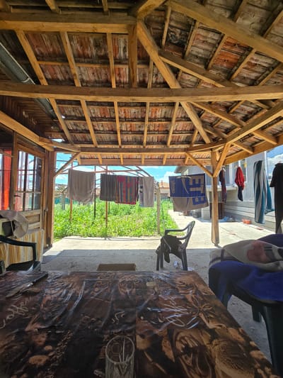 This is a wide-angle, eye-level shot of a covered outdoor patio area in Parvomay, Bulgaria. The patio has a wooden roof supported by wooden beams, with terracotta tiles visible on the underside. Towels and clothing are hanging on clotheslines across the patio, and a garden with green plants is visible beyond. In the foreground, a table covered with a patterned tablecloth is present, with a glass on it. To the right, chairs draped with blankets are visible. The overall scene depicts a domestic outdoor space.