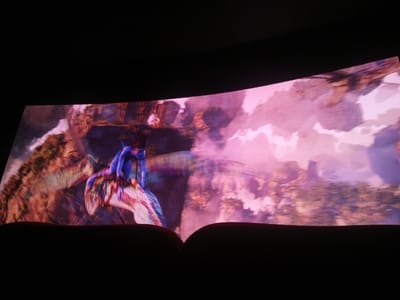 A curved cinema screen, observed from a seated position in a darkened room, displays a vibrant scene characteristic of the Avatar universe. A prominent blue, winged creature with a rider is depicted mid-flight on the left, set against a backdrop of floating landmasses and a sky dominated by pink and purple clouds. The lower foreground is obscured by dark, out-of-focus shapes consistent with the top contours of two theater seats. This viewing event is occurring within the city of Cianjur, Indonesia.
