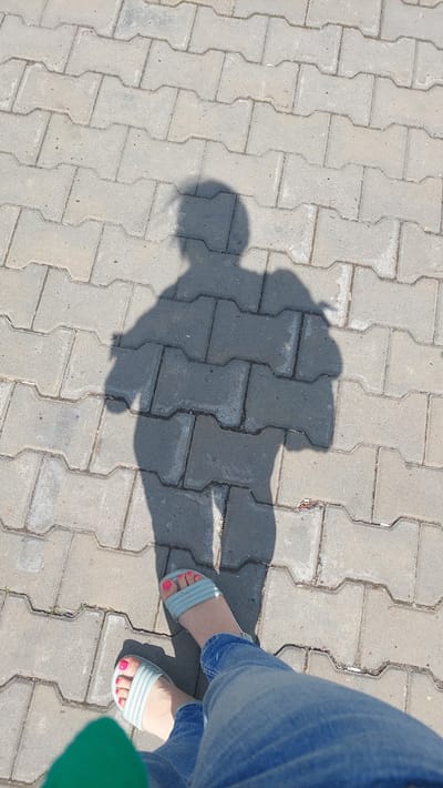 A shadow of a person, likely a woman, is cast on a paved walkway composed of interlocking concrete blocks. The shadow shows a head, shoulders, arms, and legs. The person is wearing light blue sandals with a strap across the toes, and blue denim jeans. The person's feet are visible in the foreground, with brightly painted pink toenails. A portion of a green garment is visible in the bottom left corner. The scene appears to be outdoors in daylight.