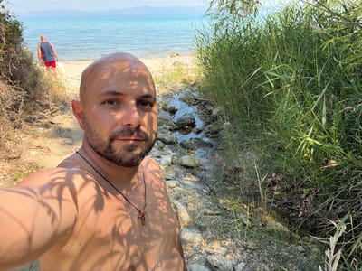 The image is a selfie taken outdoors by a man standing near a body of water. He is wearing a necklace with a wooden cross. The man is shirtless and his skin is lit by dappled sunlight, creating shadows on his chest and face. He has a beard and short hair. The background shows a rocky shoreline with sparse vegetation. In the distance, another person is visible walking away from the camera, wearing red shorts and a dark shirt. The water appears to be clear and blue, suggesting a sunny day and a coastal or lakeside setting. The image is oriented sideways, with the man's head pointing towards the bottom left of the frame. The overall impression is of a relaxed, possibly vacation setting. The location context indicates the photo was taken in Kassandra Municipal Unit, Greece. No visible text is present in the image.