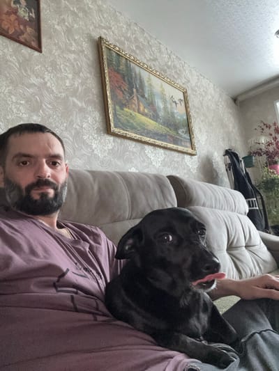 An indoor scene, likely a living room in the city of Reftinsky, Russia, shows a male individual and a black dog seated on a light-colored upholstered sofa. The man, positioned on the left, has dark hair and a beard, wears a purple t-shirt, and looks directly at the camera. To his right, a black dog sits with its head turned right, eyes wide open, and its tongue slightly extended from its mouth. The wall behind them is covered in light-toned patterned wallpaper. A large, ornate gold-framed landscape painting depicting a cabin, trees, water, and birds in flight hangs prominently on the wall. A smaller, darker framed image is partially visible in the upper left. In the upper right background, a dark object resembling a bag, a white decorative birdcage, and several potted plants are visible.